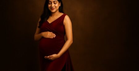 Maternity photoshoot in Gurgaon featuring a South Asian pregnant woman in a red velvet gown, softly lit against a warm brown backdrop, cradling her baby bump with a serene smile – Baby Bum Photography.