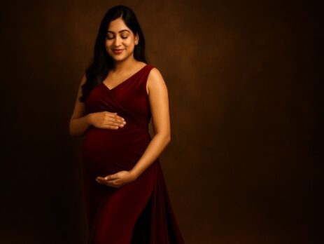 Maternity photoshoot in Gurgaon featuring a South Asian pregnant woman in a red velvet gown, softly lit against a warm brown backdrop, cradling her baby bump with a serene smile – Baby Bum Photography.